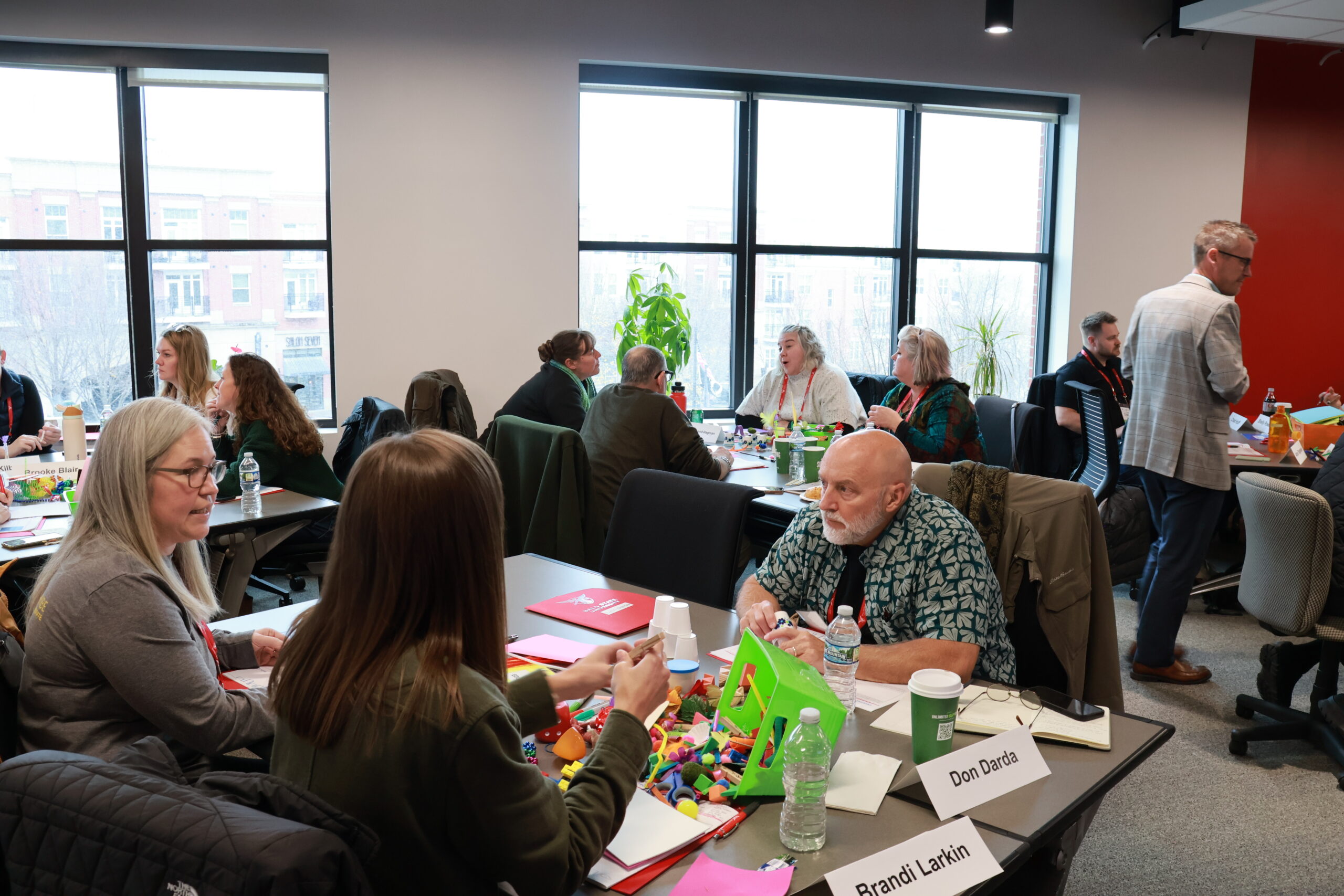 Three adults sitting at a conference table among other small groups at other tables. An overturned bin of craft supplies is spilling onto the table between them.