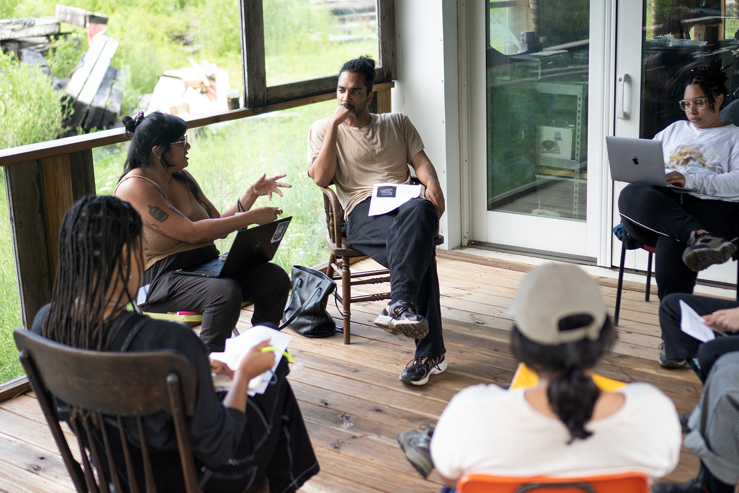 A group of people sitting on chairs in a circle as one person gestures and talks. Some have sheets on paper on their lap, while others take notes on their laptops.