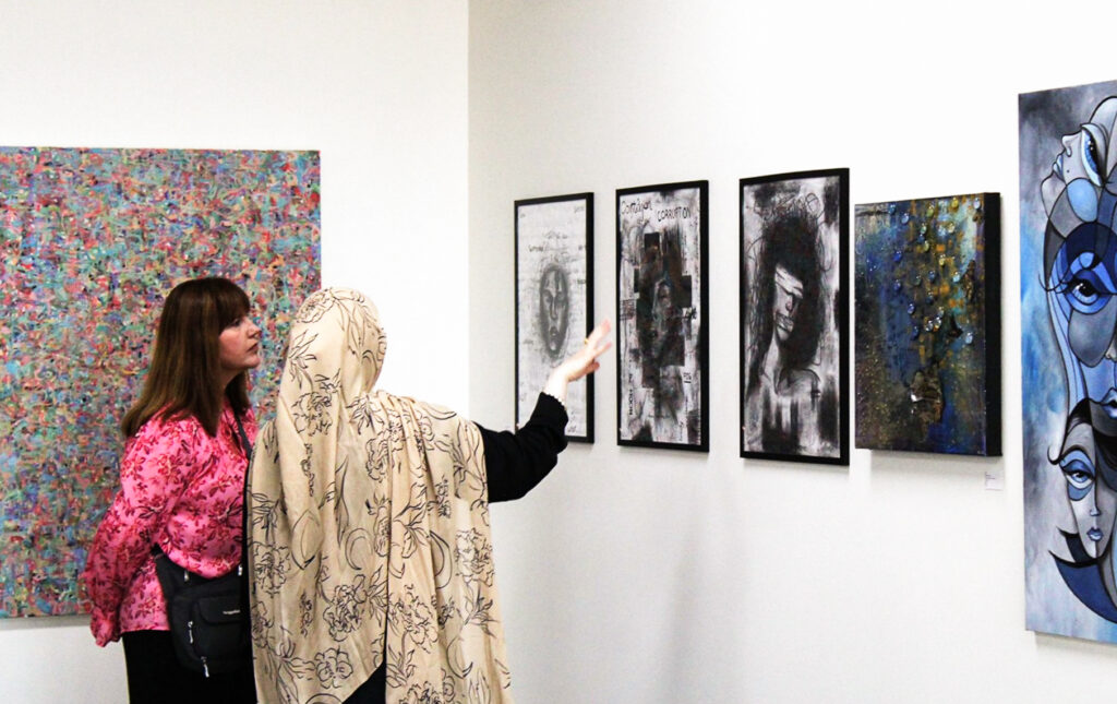 Two people standing together by a wall of paintings. One person is gesturing towards a painting as the other looks on.