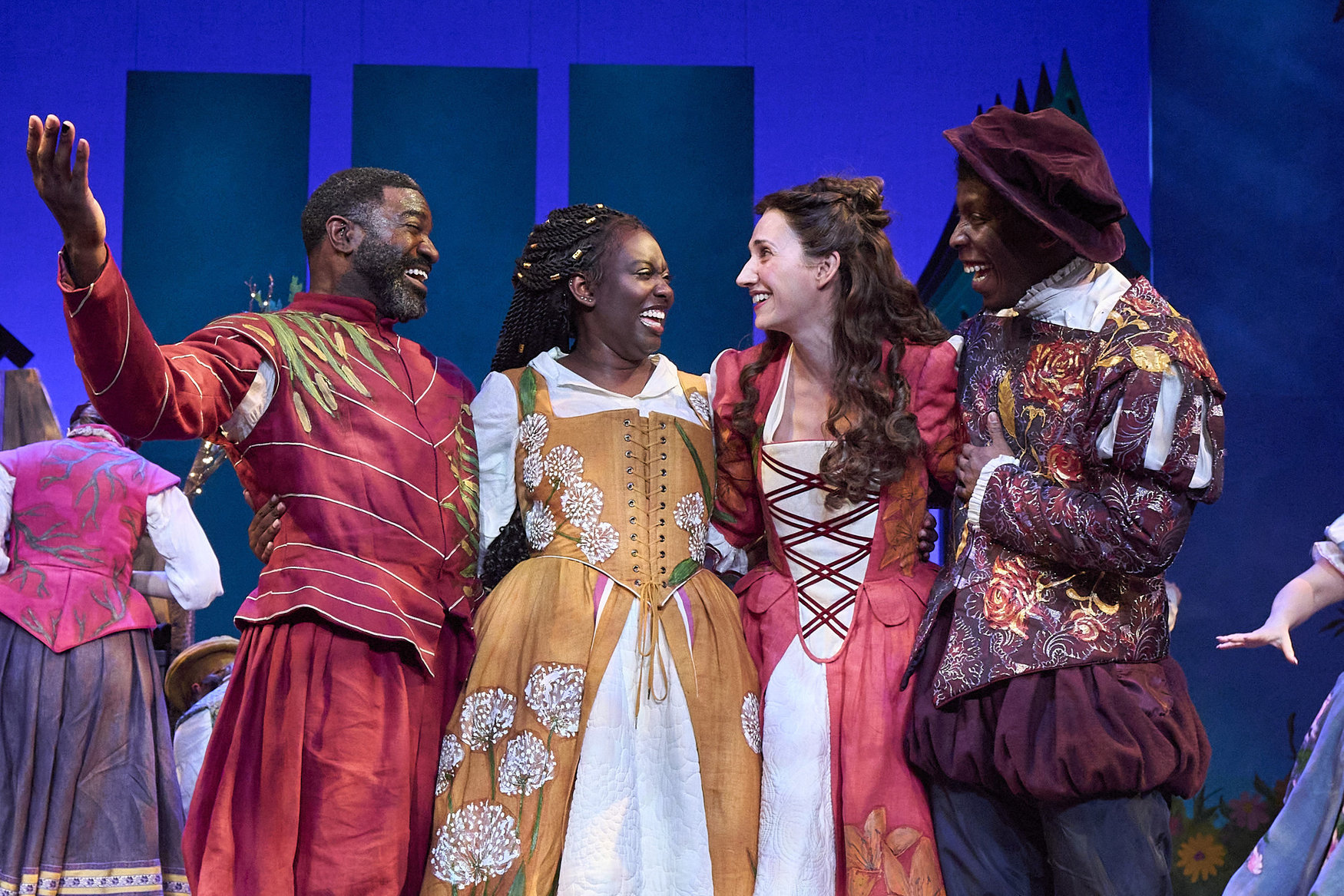 A group of four smiling people with arms around each other, dressed in period costumes on a stage.