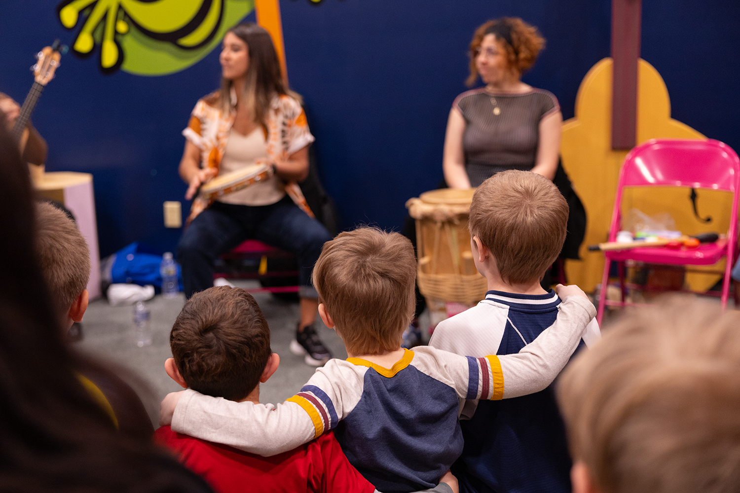 A group of young children sits closely together, two with their arms around each other, watching musicians play hand drums in a brightly colored playroom.