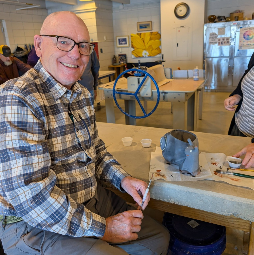 A man with light skin and a flannel shirt holds a paintbrush and poses next to a snail planter.