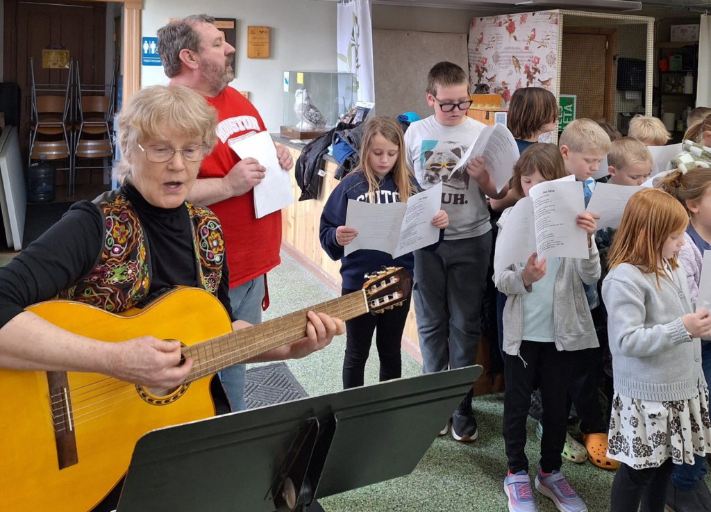 A dozen or do children holding choral music and singing next to a guitarist.
